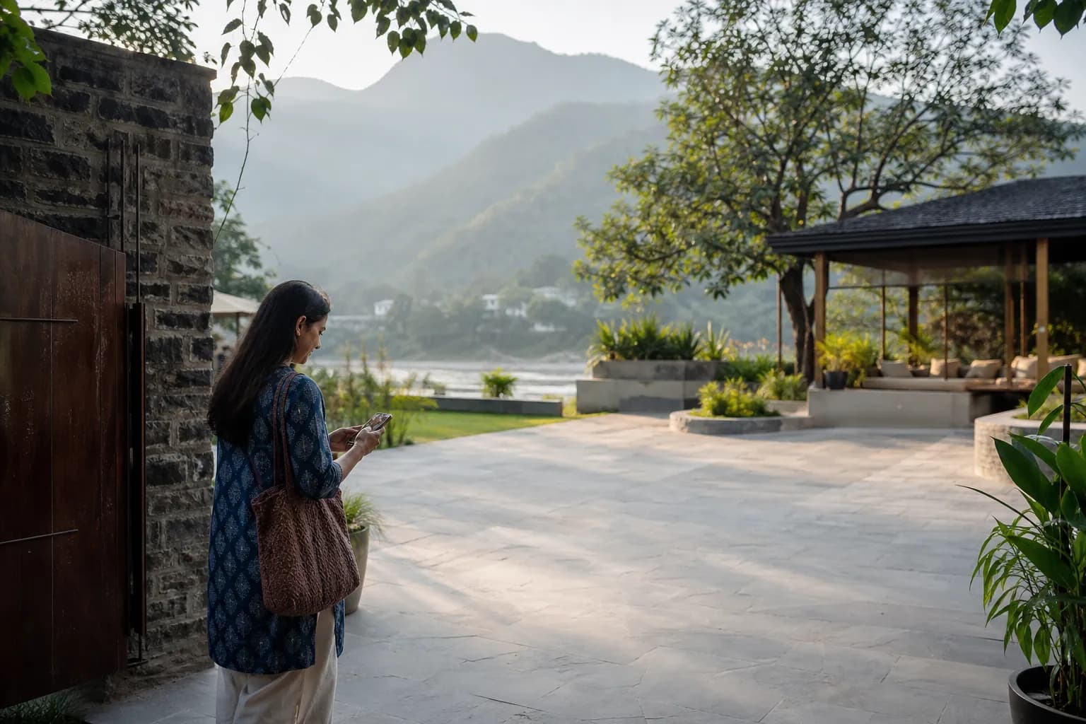 A woman checking her phone in a quiet riverside courtyard beneath forested hills